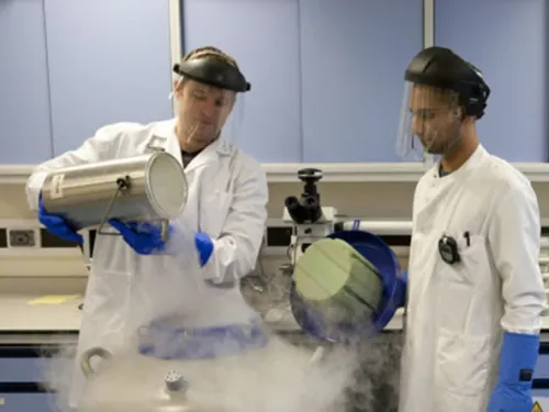 Stuart and Ruman in the lab, wearing white coats and face shields whilst pouring liquid nitrogen.