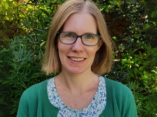 A woman with shoulder-length hair and glasses is standing in front of a green hedge. She is wearing a floral T-shirt and a green cardigan. 