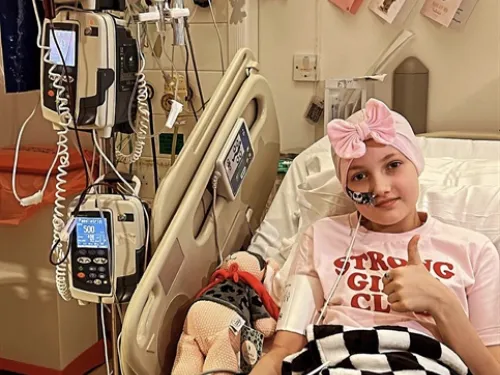 A young girl wearing a pink headband and a t-shirt that says "Strong Girls Club" is lying in a hospital bed surrounded by medical equipment.