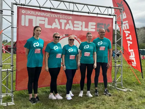 A group of five individuals, consisting of four women and one man, stands together in front of a red inflatable background marking the start of a 5K race. They are all wearing matching teal t-shirts with the CCLG logo.