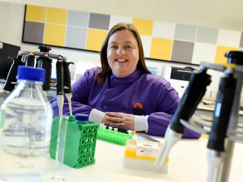 Vikki, smiling at the camera and wearing a purple lab coat whilst surrounded by lab equiptment.