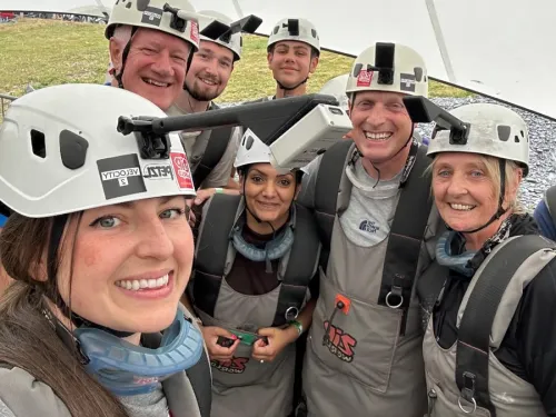 A group of 6 smiling people, all wearing helmets and safety gear for doing a zip line challenge, are taking a group selfie.