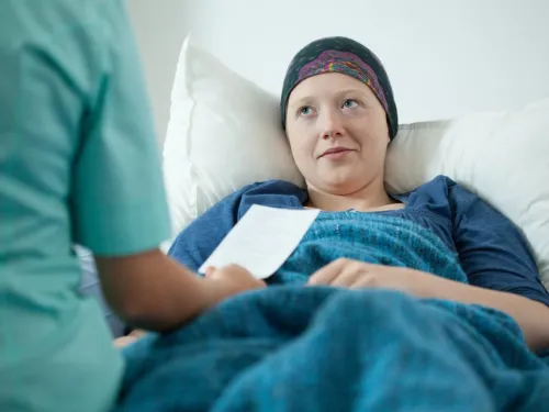 A young woman wearing a headscarf, lying under a blanket, talking to a doctor.