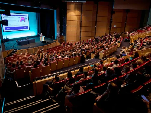 People sitting in a large room, watching someone give a presentation on a stage.