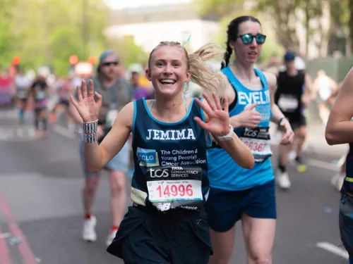 A woman with blonde hair, running in a CCLG running top whilst smiling and waving at the camera