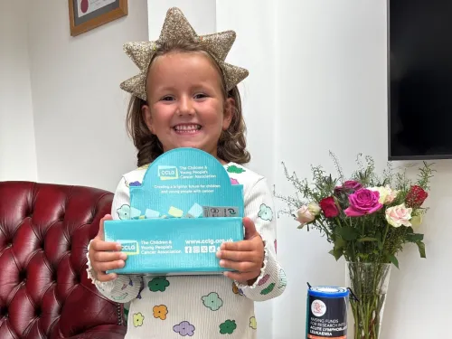 A young girl in spotty pyjamas wearing a star headband, smiling and holding a CCLG badge box in front of her