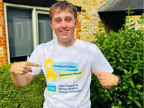 A young man smiling at the camera and pointing to his t-shirt, which is white with the CCLG and Childhood Cancer Awareness Month logos, 
