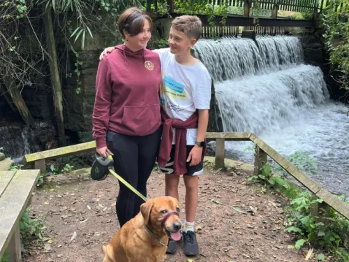 A mum and son smiling and hugging in front of an outdoor water feature, holding a dog on a lead