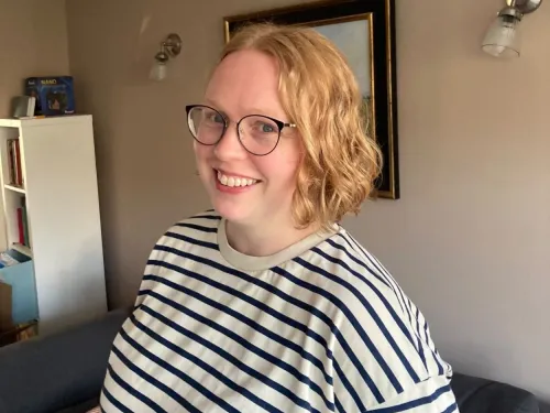 Anna has a blonde bob and is smiling at the camera indoors, wearing a stripey shirt and glasses