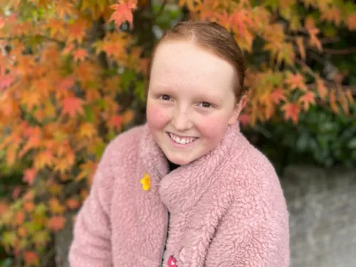 Young girl smiling at camera with pink coat and ginger hair standing in a park with autumn leaves in the bakground