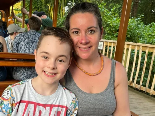 A boy wearing a marvel t shirt and his mum wearing a grey vest and orange necklace sitting in a train