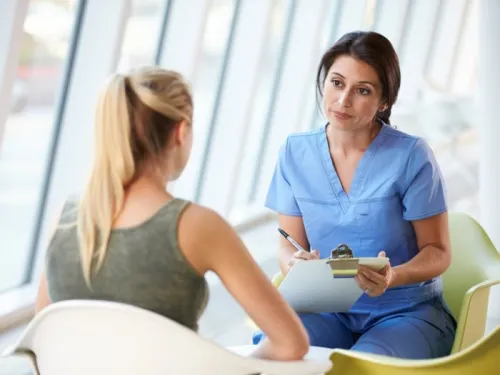 diverse nurse with olive skin holding a clipboard talking to teenager wearing a vest top  with blonde hair in a pony tail